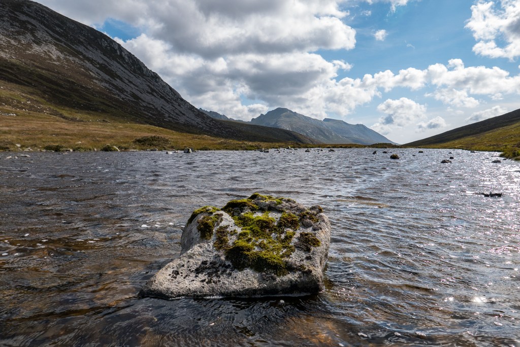 Shallow loch with a mossy rock in the water, mountains in the distance