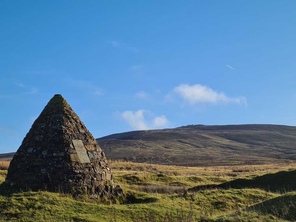 Pyramid shaped cairn on the left built with stones, hills, blue sky and white clouds in the background