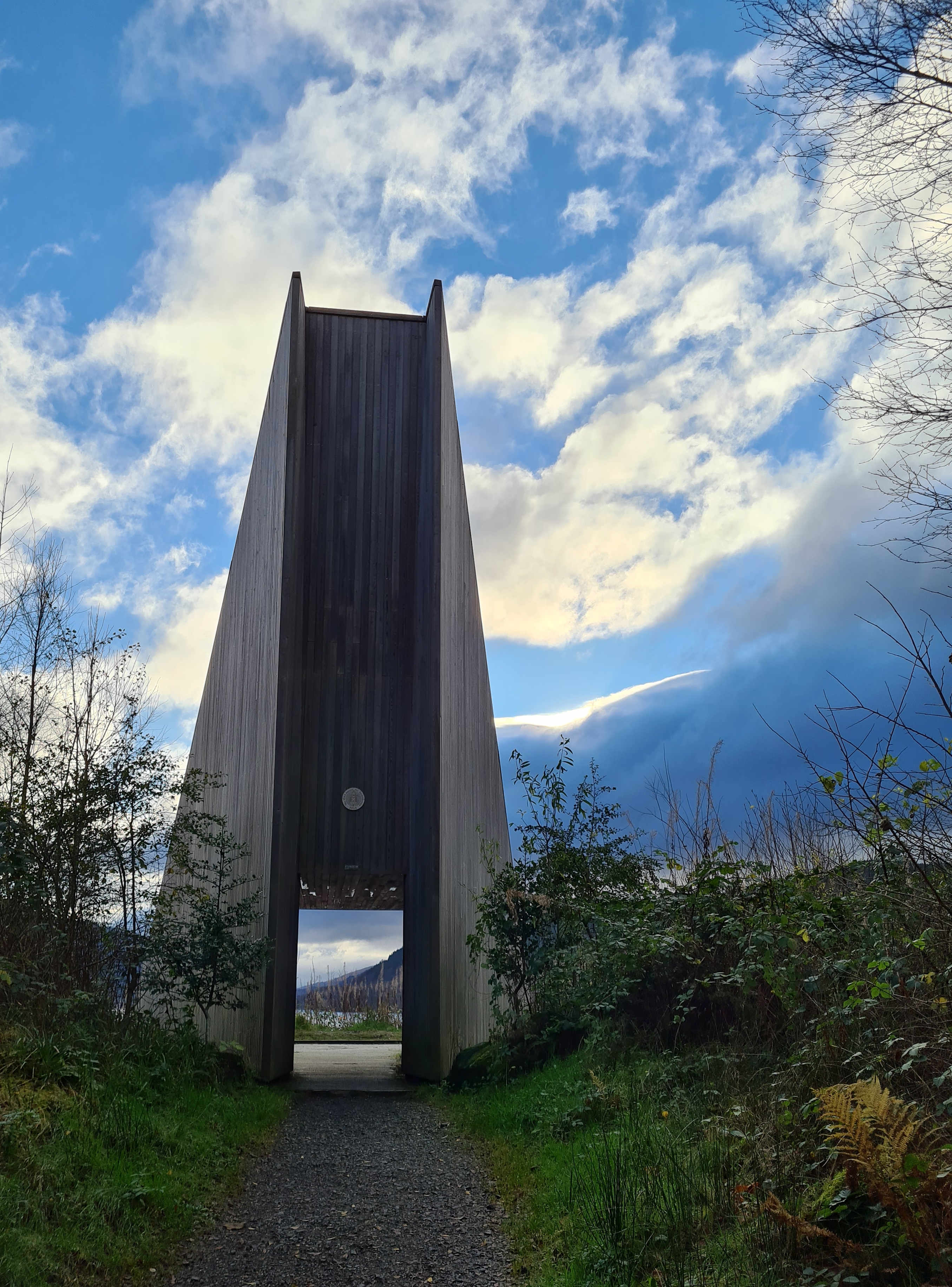 Wooden pyramid like shape structure with an open entrance at the bottom, blue sky and white puffy clouds