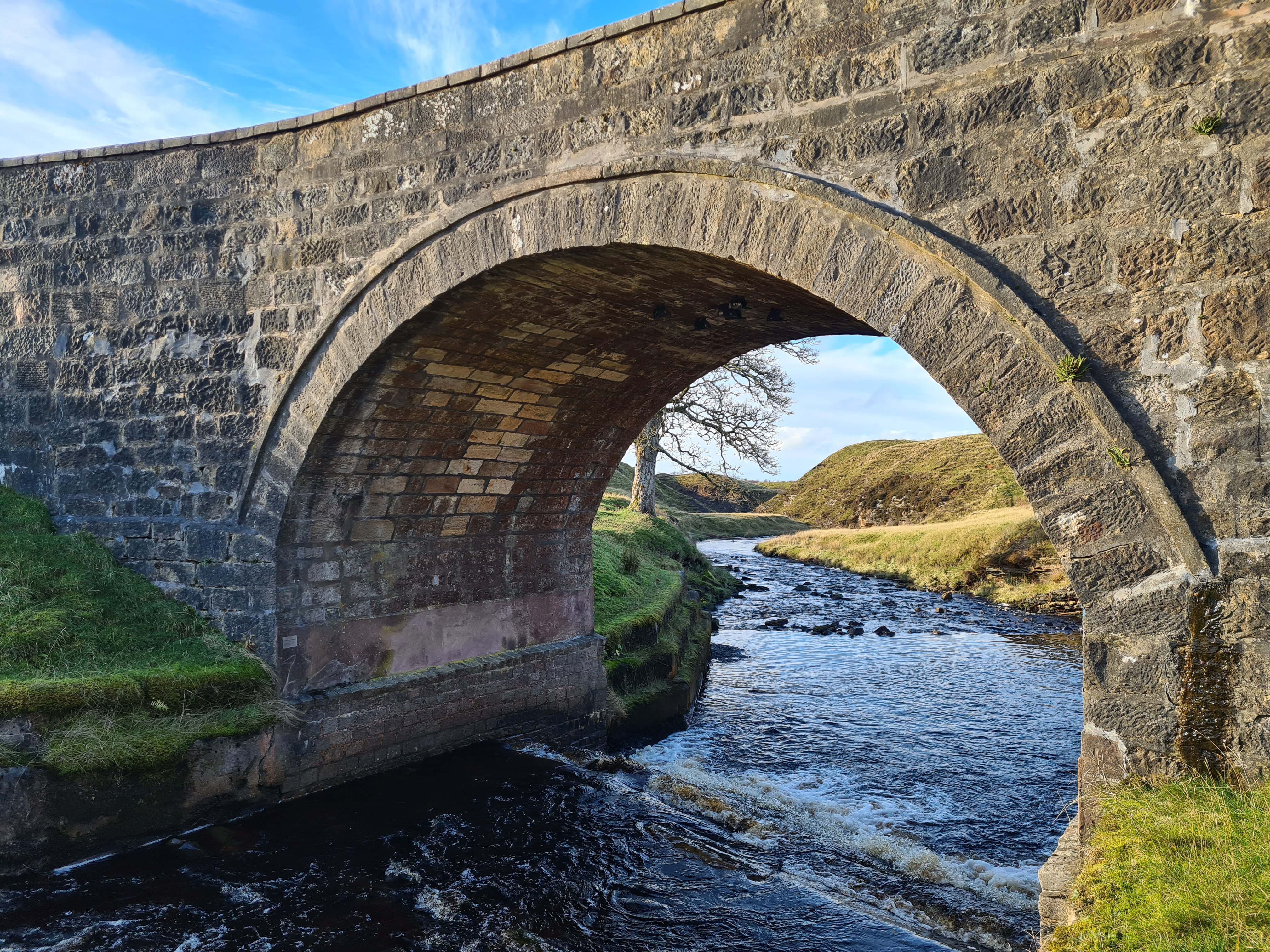 Looking through the arch of an old stone bridge, water flowing through, tree on the banks of the water