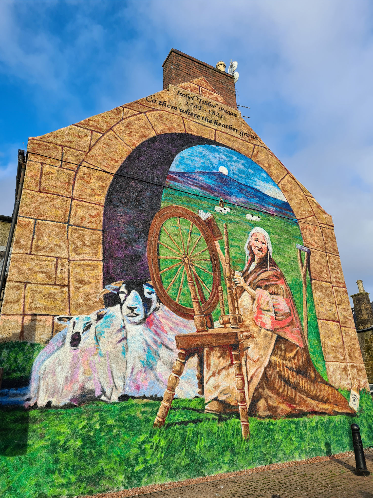 Mural painting of an elderly lady sitting at a spinning wheel, beside a bridge arch and sheep
