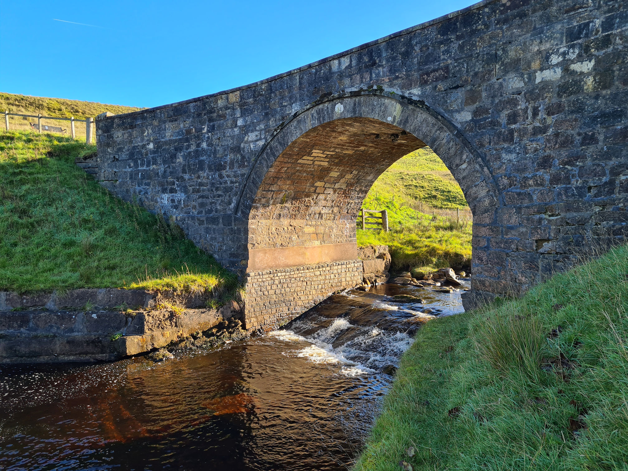 The arch of a stone bridge with water flowing underneath like a small waterfall. The sunlight in shining under the arch and on the water.