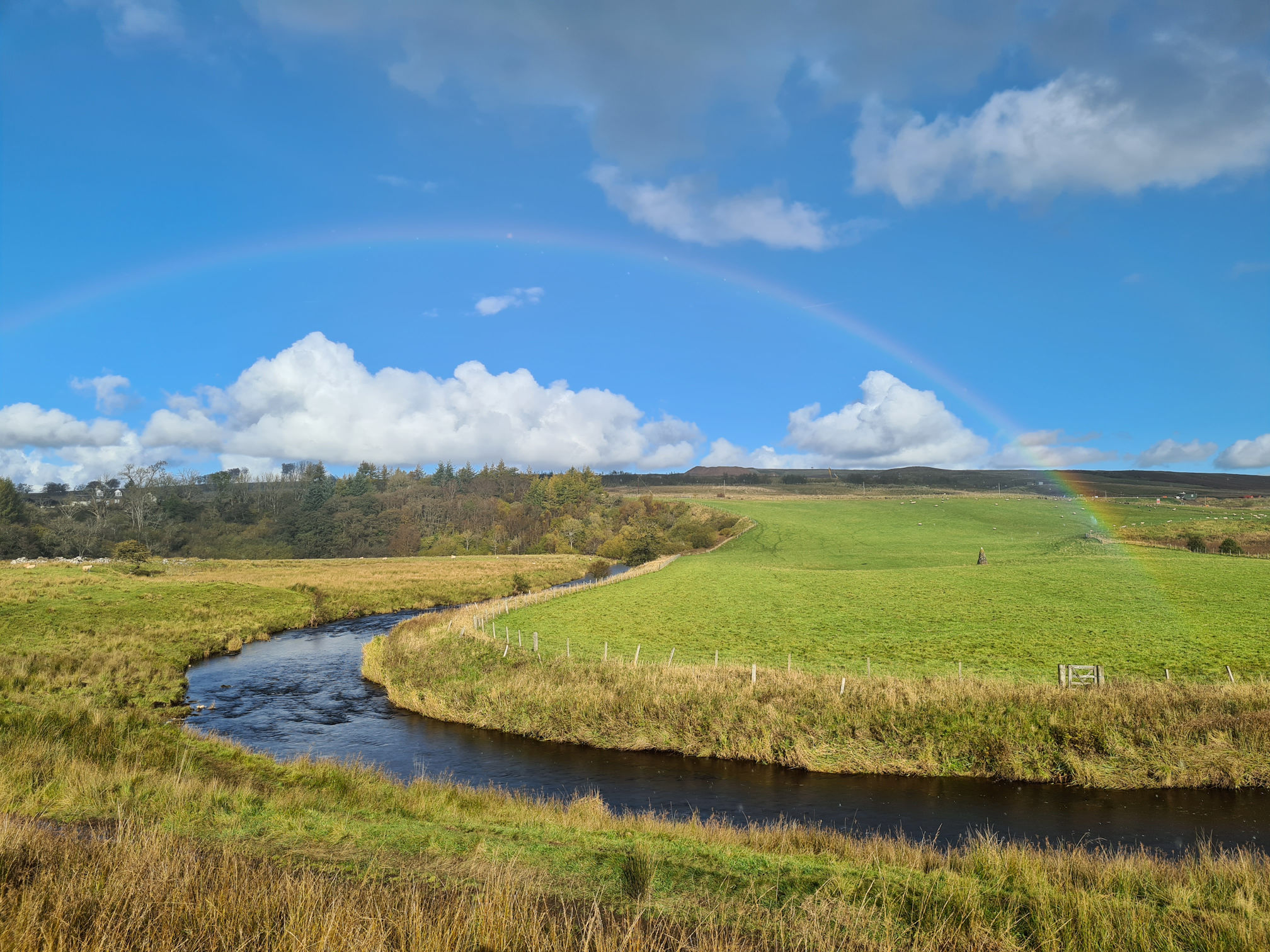 A beautiful rainbow arches over the meandering river, bright blue sky, puffy white clouds, green and brown fields framing the river