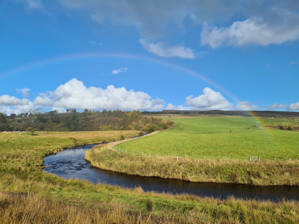A beautiful rainbow arches over the meandering river, bright blue sky, puffy white clouds, green and brown fields framing the river