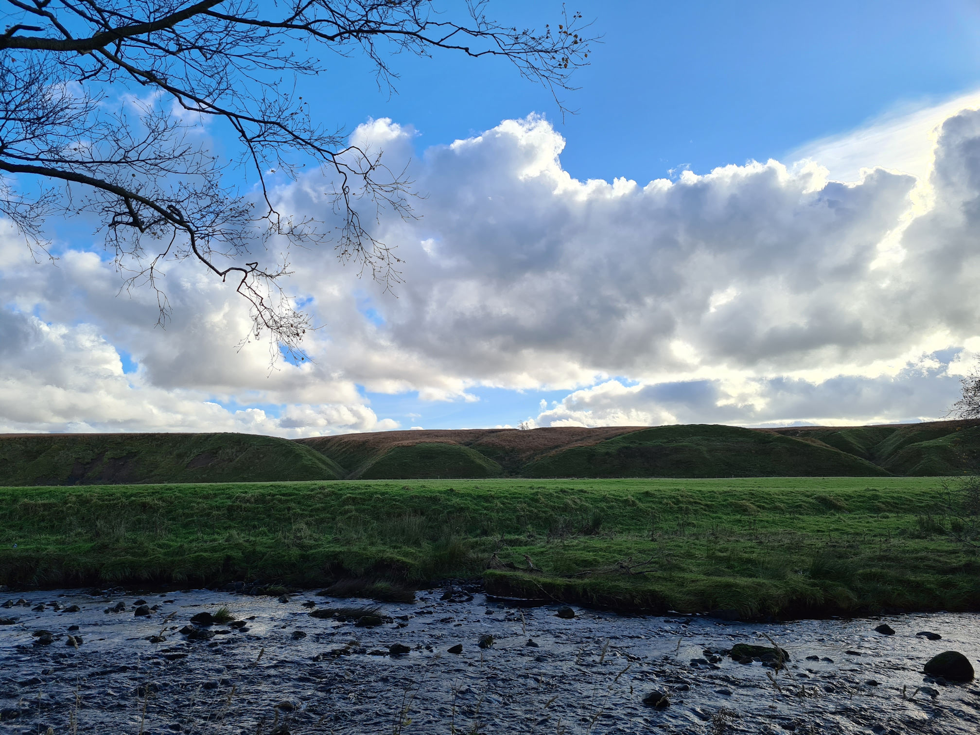 River, sun is hiding behind white puffy clouds making the light dark, small hill mounds across the water