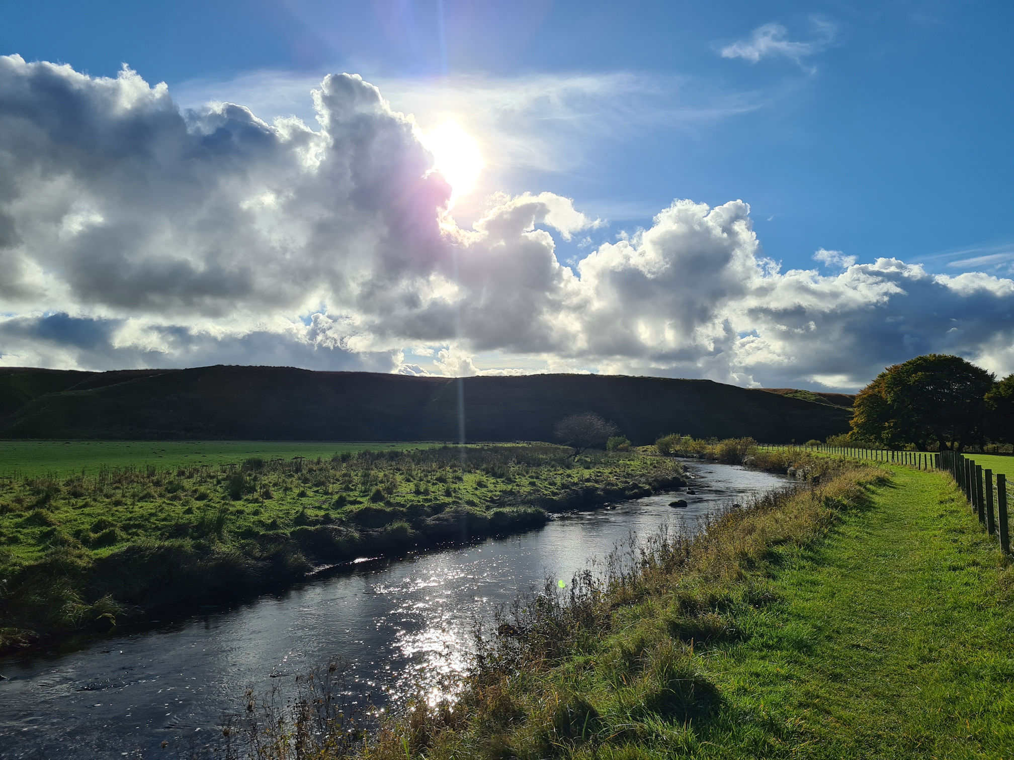 River, green grassy path, large puffy white clouds, the sun is peaking through
