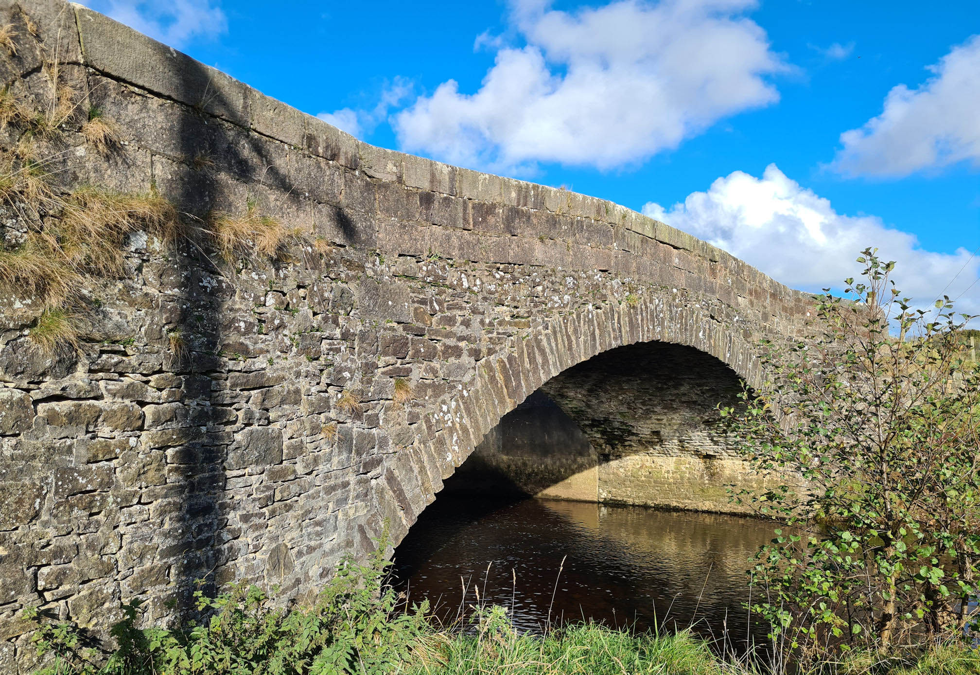 Old stone bridge, water flowing underneath, sunlight is shimmering under the arch