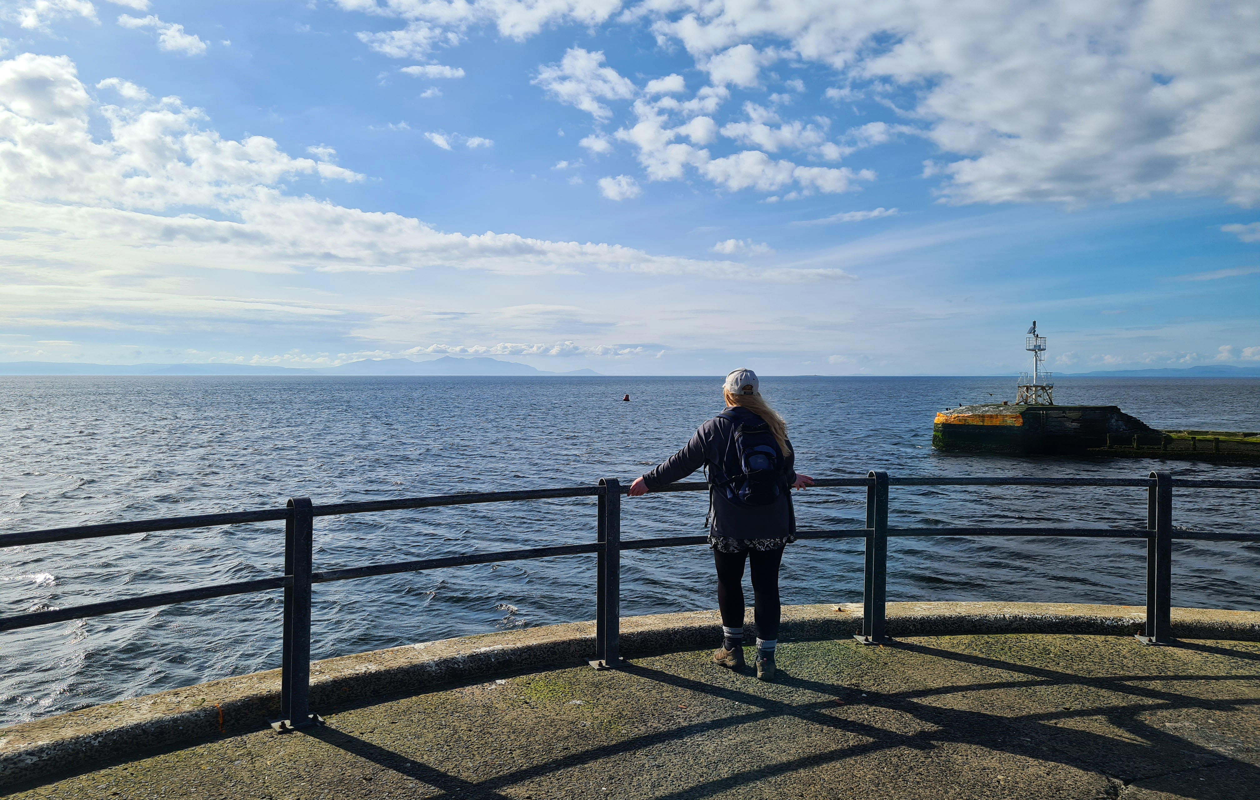 Girl standing at the end of a harbour, holding on to a railing and looking out to sea at the island far away in the distance, wishing she was there