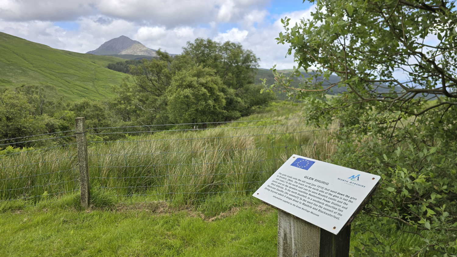 Information sign about the old road of Glen Shurig, with a view across open grassy fields looking towards the mountain peak Goat Fell