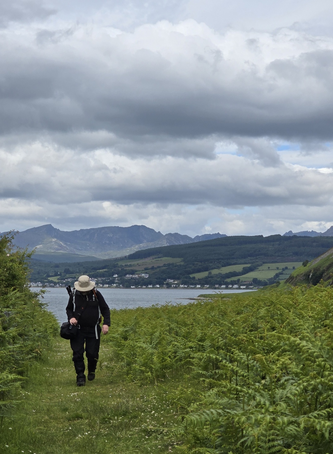 A man with a hat walking on a grassy path, head down, mountains and sea in the background