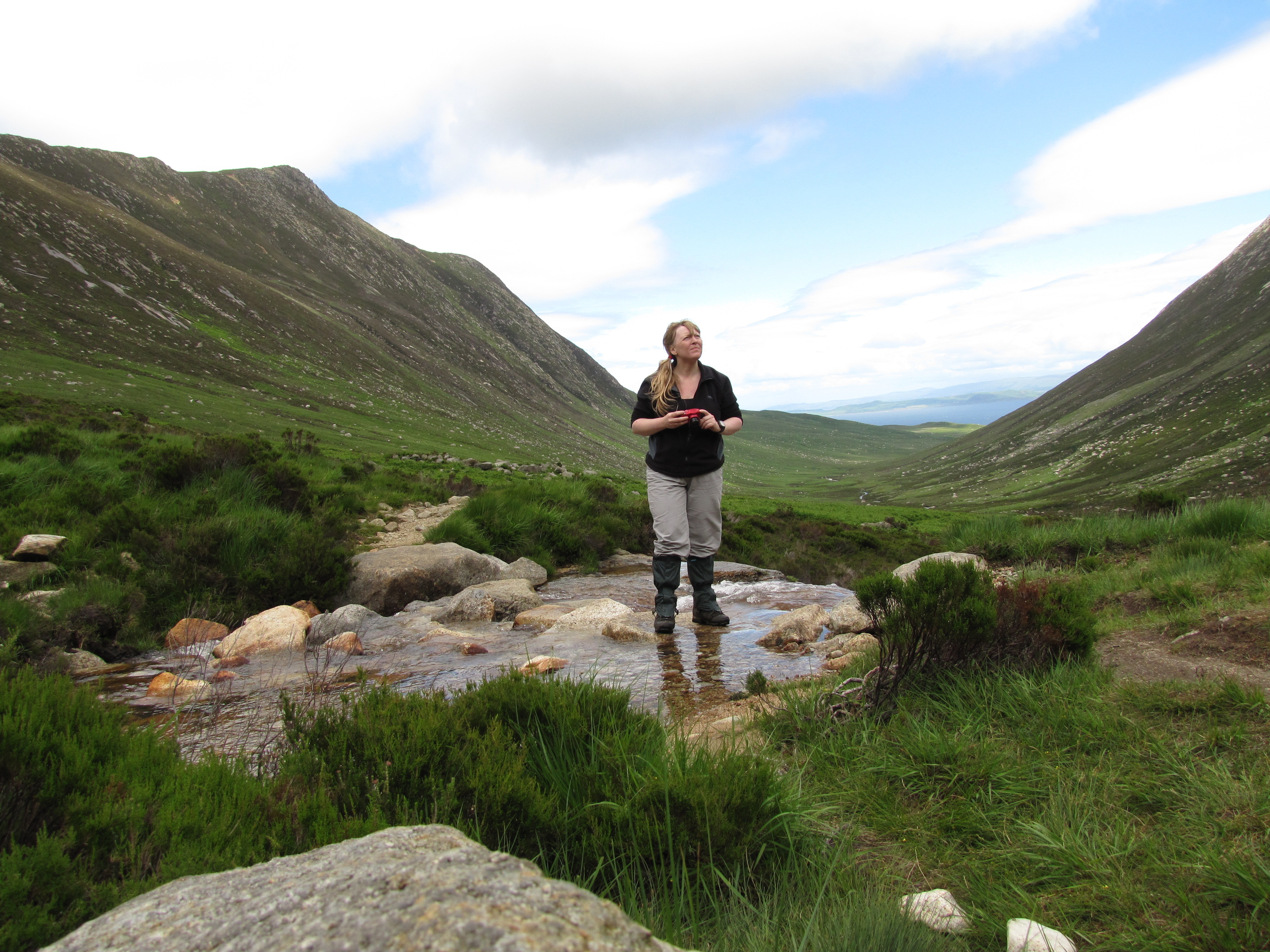 Girl standing in the Sannox Burn with a camera in her hand and looking around at the mountain views