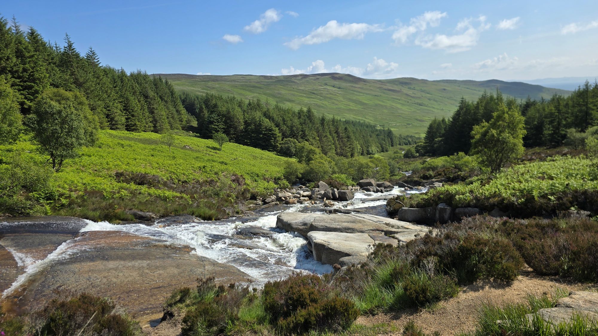 Water falling down a rocky burn, trees and hills surround the burn