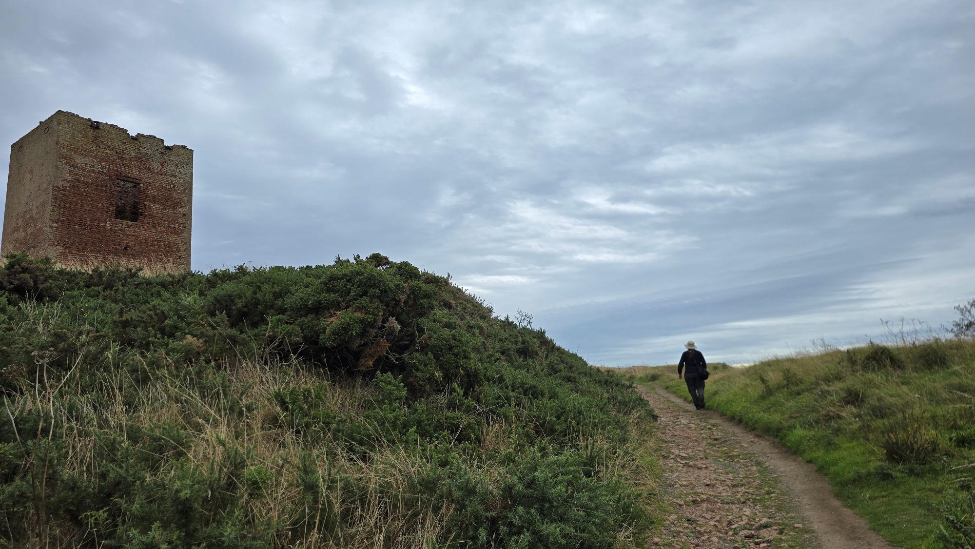 Person walking on a rough path towards the coast with a small red brick dovecot ruin on the left